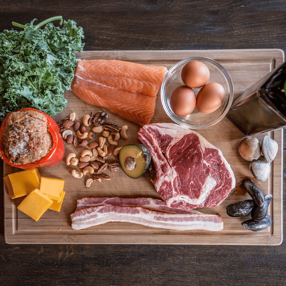 Whole foods laid out on a cutting board: salmon, eggs, steak, kale, nuts, avocado and cheese